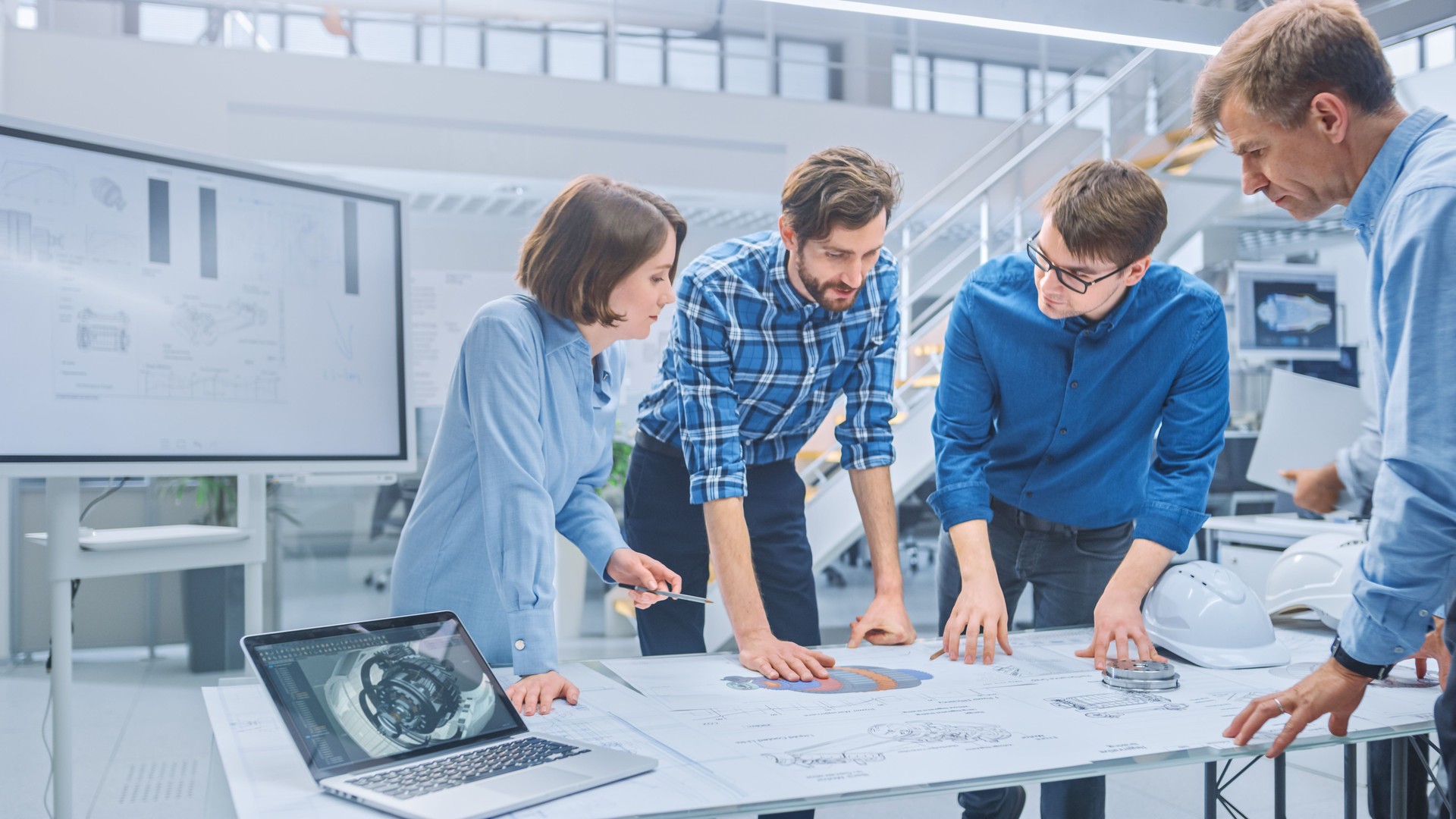 In the Industrial Engineering Facility: Diverse Group of Engineers and Technicians on a Meeting Gather Around Table with Engine Design Technical Drafts, Have Discussion, Analyse Technology